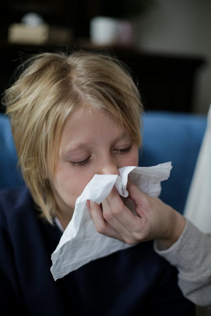 sick-child-wiping-his-nose-with-tissue-5858852 Young girl sneezing into a tissue while being comforted. Indoor setting.